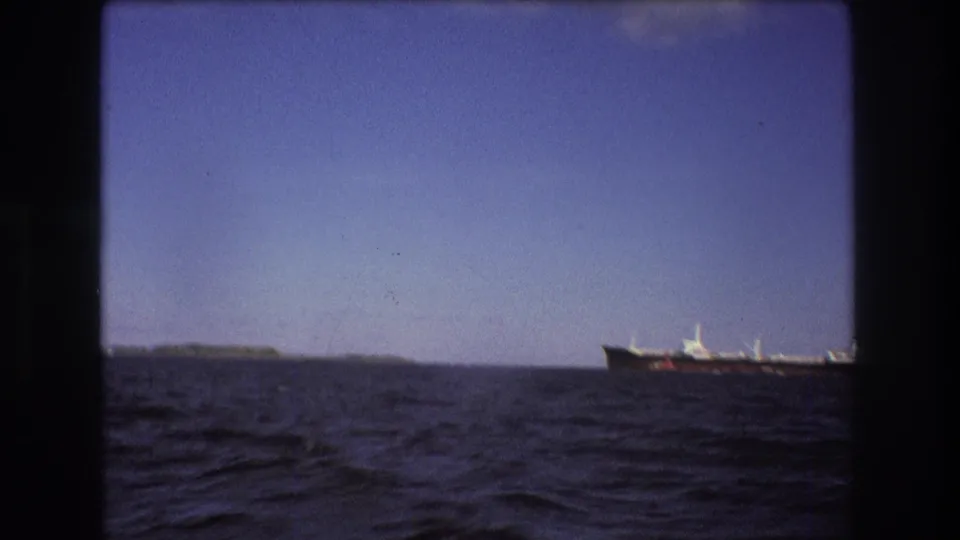 1975 Boston-area 8mm frame showing a distant large ship crossing the horizon beyond choppy open water and a low shoreline.