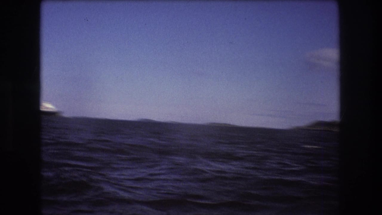 1975 Boston-area 8mm frame with rolling ocean water, a low dark shoreline, and blue sky seen through a dark film gate border.