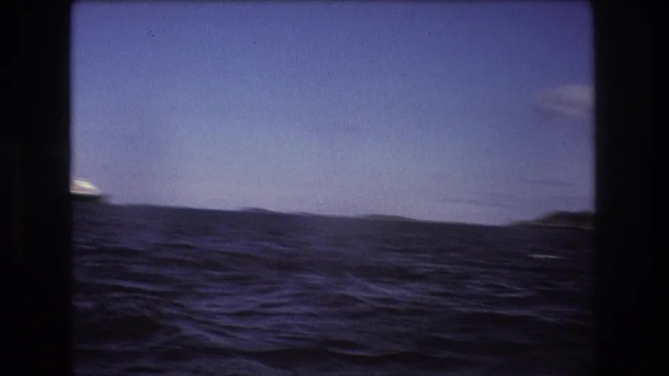 1975 Boston-area 8mm frame with rolling ocean water, a low dark shoreline, and blue sky seen through a dark film gate border.