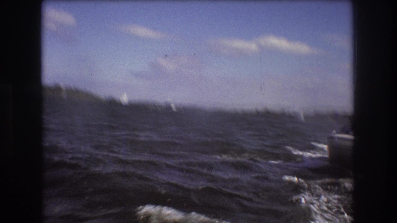 1975 Boston-area 8mm frame looking across choppy water toward a low shore, with small bright sail-like shapes visible in the distance.