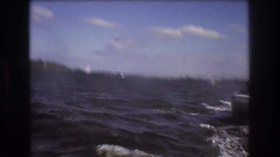 1975 Boston-area 8mm frame looking across choppy water toward a low shore, with small bright sail-like shapes visible in the distance.