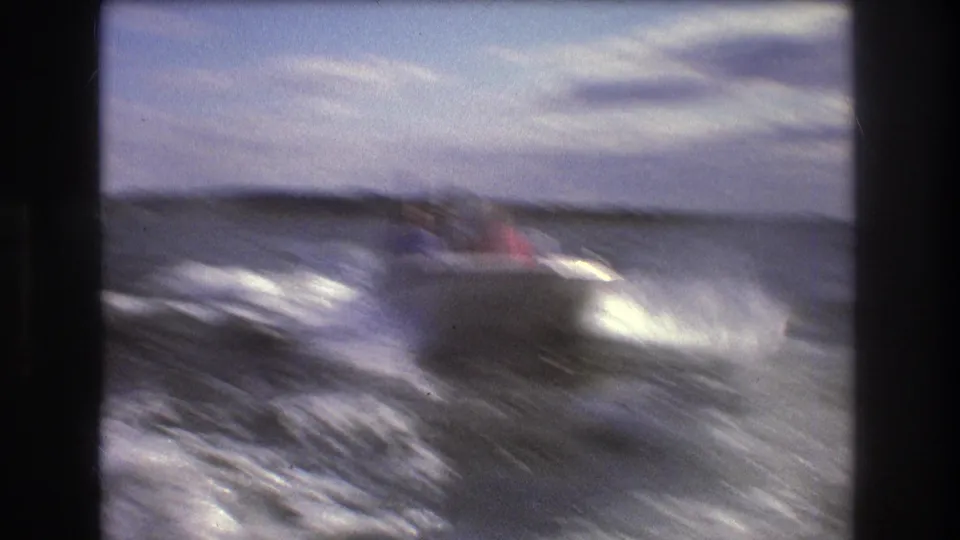 1975 Boston-area 8mm frame showing a small speedboat with passengers riding through choppy open water, with wake and spray behind it.