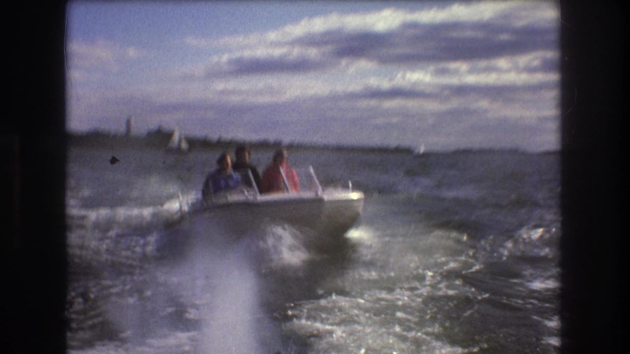 1975 Boston-area 8mm frame showing a small white motorboat with seated passengers moving through choppy harbor water, with distant sailboats and shoreline behind it.