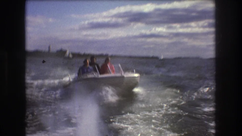 1975 Boston-area 8mm frame showing a small white motorboat with seated passengers moving through choppy harbor water, with distant sailboats and shoreline behind it.