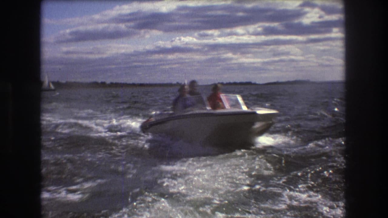 1975 Boston-area 8mm frame of a small white motorboat passing over dark, rippled harbor water with passengers visible and a low shoreline in the distance.