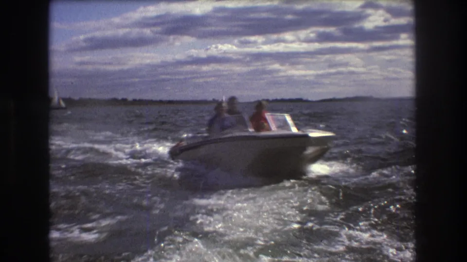 1975 Boston-area 8mm frame of a small white motorboat passing over dark, rippled harbor water with passengers visible and a low shoreline in the distance.