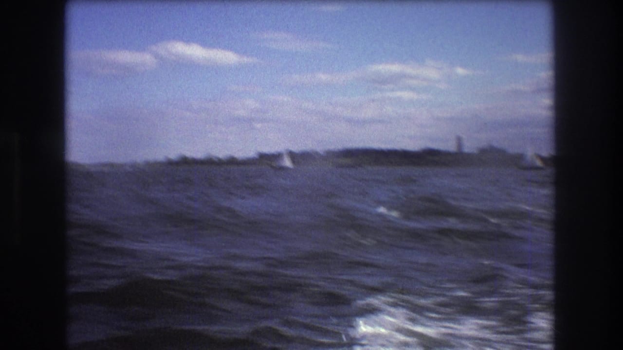 1975 Boston-area 8mm frame looking across open harbor water toward distant sailboats, a low shoreline, and a lighthouse-like vertical structure on the horizon.