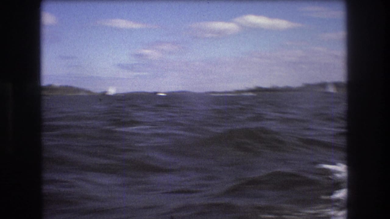 1975 Boston-area 8mm frame of rippled harbor water with distant shoreline and small sailboats under a pale blue, partly cloudy sky.