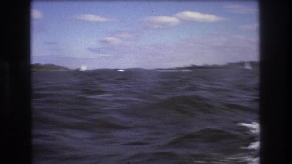 1975 Boston-area 8mm frame of rippled harbor water with distant shoreline and small sailboats under a pale blue, partly cloudy sky.