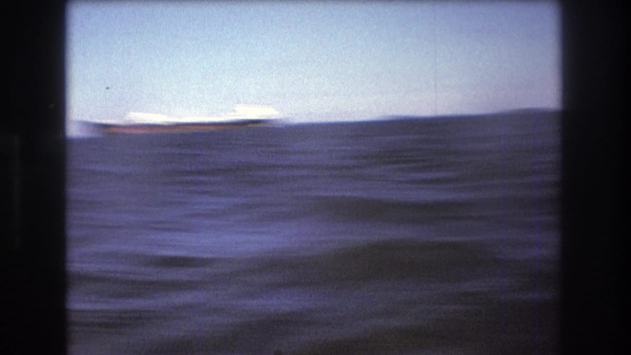 1975 Boston-area 8mm home-movie frame showing rough open water with a low distant shoreline and pale sky, filmed from a moving boat.