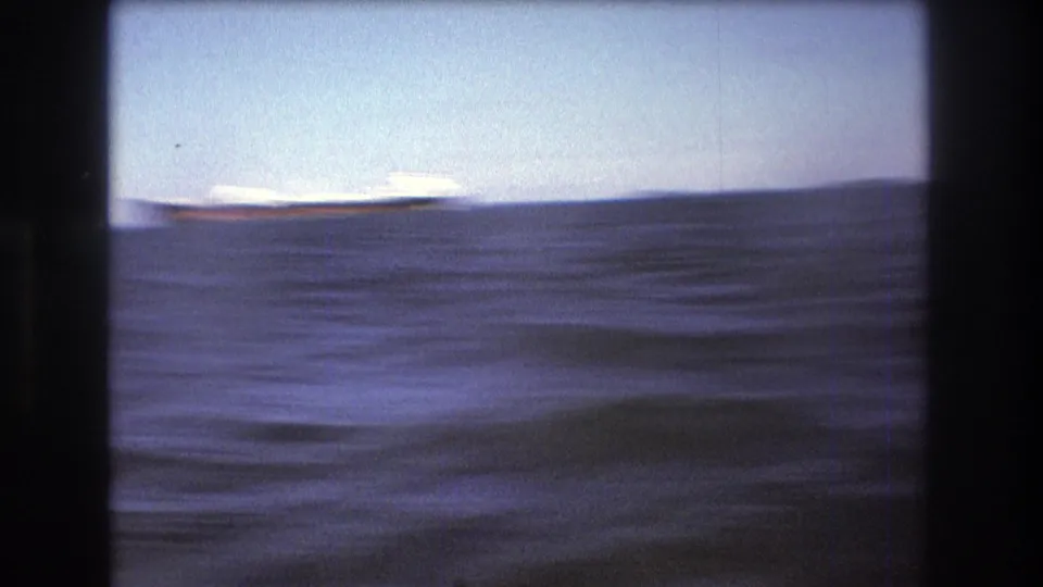 1975 Boston-area 8mm home-movie frame showing rough open water with a low distant shoreline and pale sky, filmed from a moving boat.
