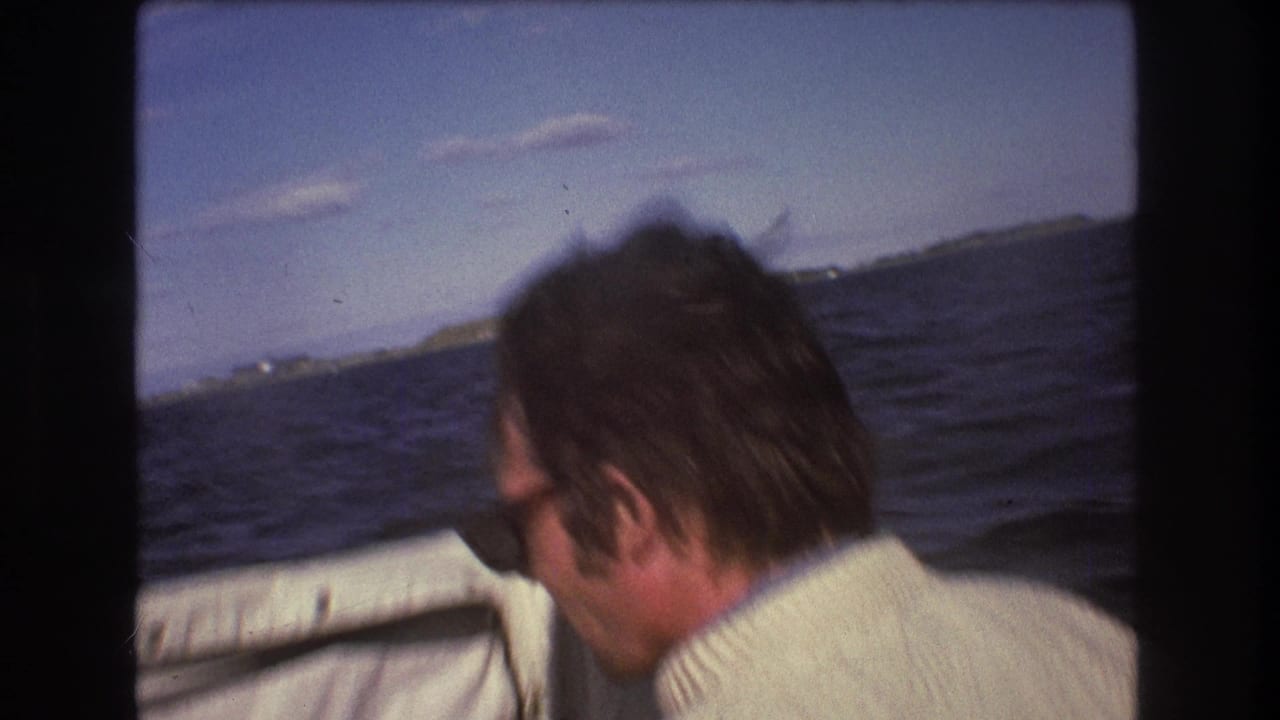 Dark-haired adult man wearing sunglasses and a light sweater looks across choppy water from inside a small boat in 1975 Boston-area home-movie footage.