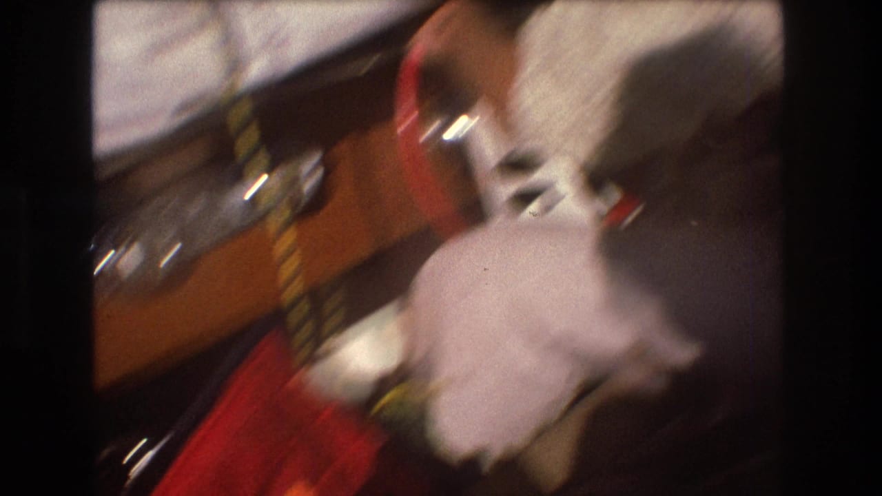 Shaky 8mm frame from inside the boat showing a blurred adult figure, wooden trim, red fabric, and onboard objects as the camera moves with the water.