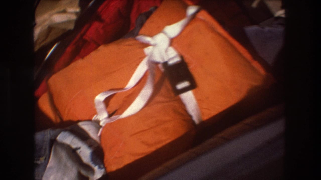 Close handheld 8mm view of an orange duffel bag with white straps beside a crumpled paper bag and surrounding travel items in the 1975 Boston home-movie clip.