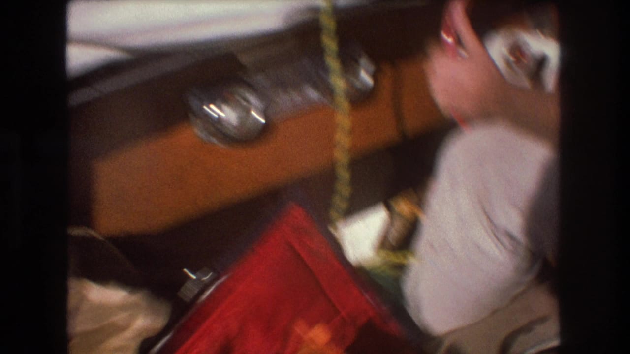 Handheld 8mm frame showing packed red fabric, a metal fitting, rope or cord, and a partial adult figure among travel items during the Boston boating outing.