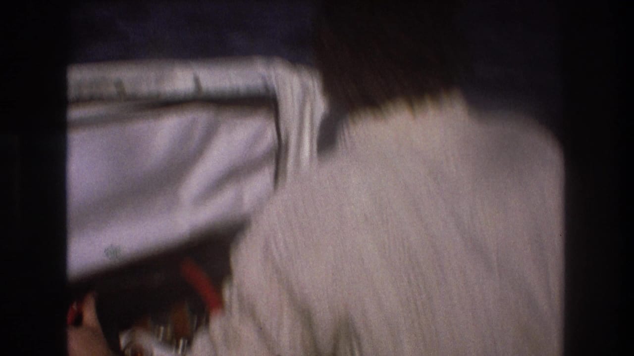 Rear view of an adult man in a white knit sweater looking forward past a boat windshield toward the water in the 1975 Boston home-movie footage.