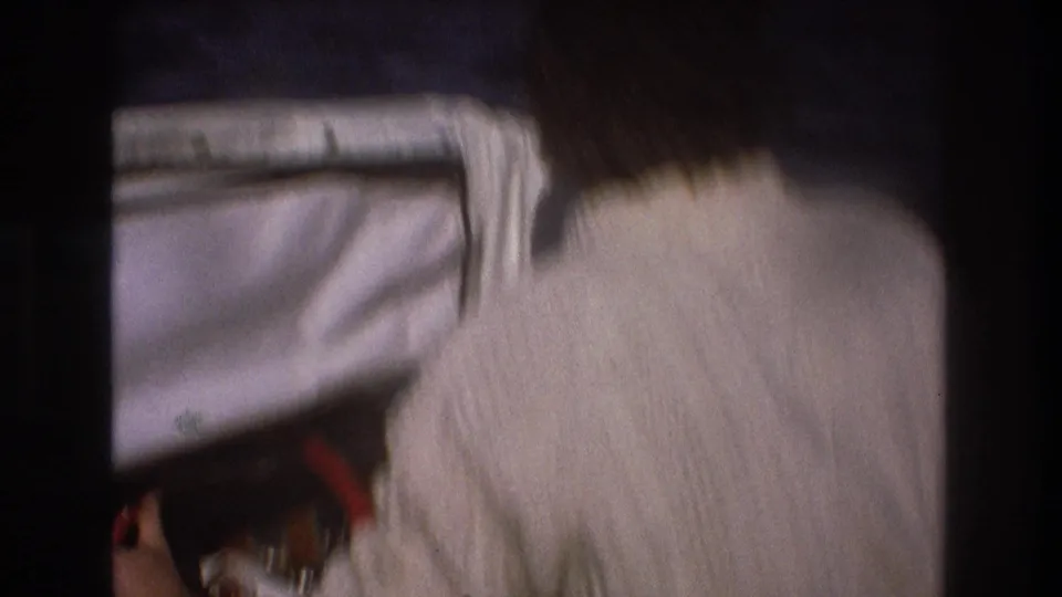 Rear view of an adult man in a white knit sweater looking forward past a boat windshield toward the water in the 1975 Boston home-movie footage.