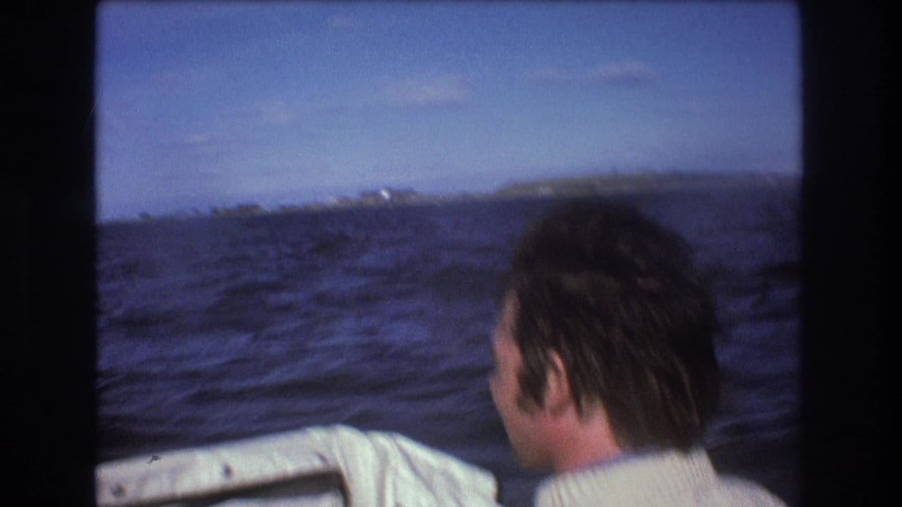 1975 Boston-area 8mm home-movie frame showing a man in a white sweater seen from behind on a moving boat, looking across choppy blue water toward a low shoreline.