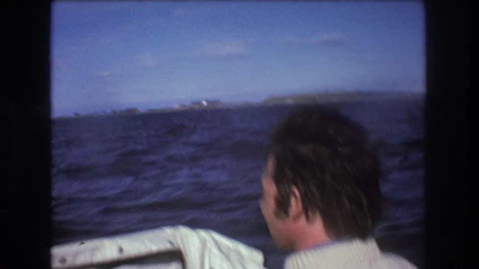 1975 Boston-area 8mm home-movie frame showing a man in a white sweater seen from behind on a moving boat, looking across choppy blue water toward a low shoreline.