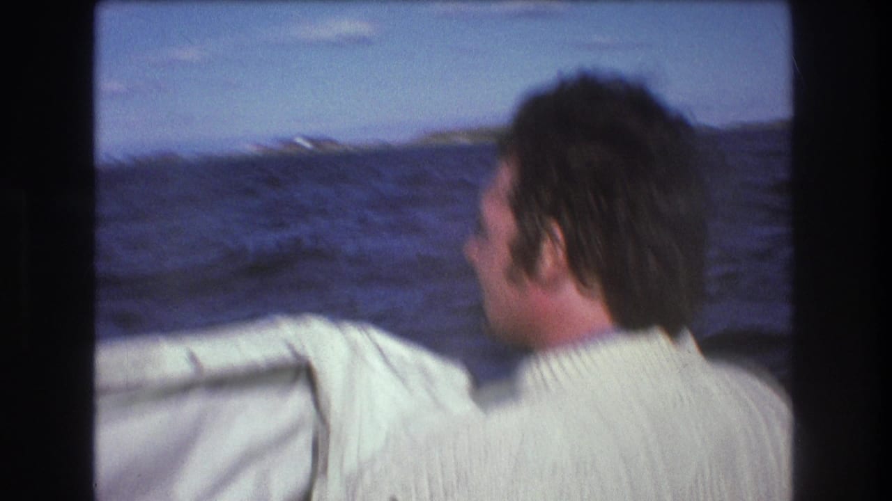 Man in a white sweater faces the water from a fast boat with shoreline in the distance.