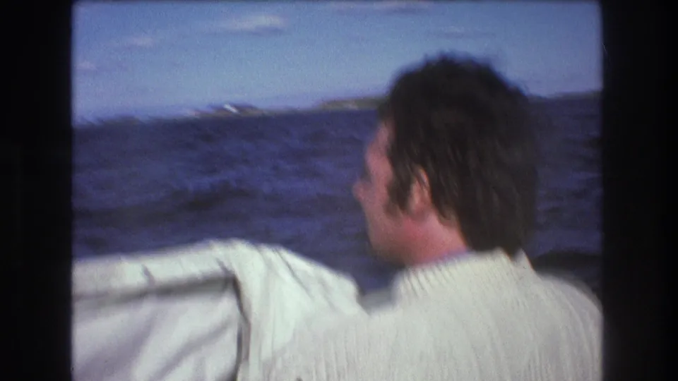 1975 Boston-area home-movie still of a man riding near the edge of a boat as dark blue water, a low horizon, and blurred shoreline pass behind him.