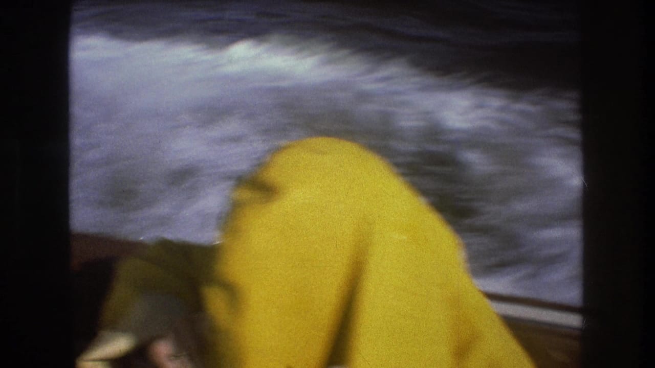 1975 Boston-area 8mm frame with a yellow hooded raincoat in the foreground, white boat spray along the side, and dark water blurred by the boat's speed.