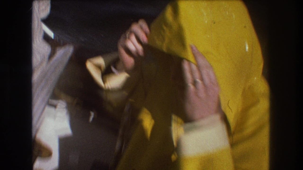 1975 Boston-area home-movie still showing hands pulling on a bright yellow hooded raincoat aboard a moving boat, with the boat interior and dark water partly visible.
