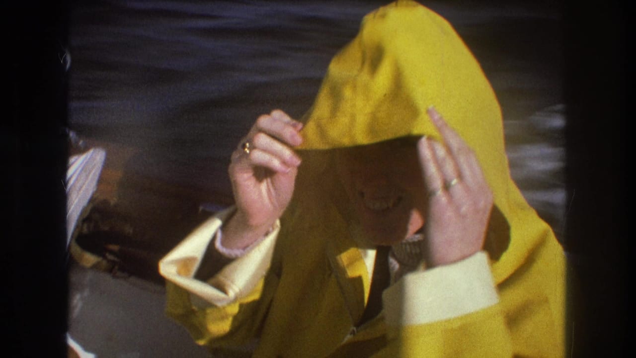 In 1975 Boston-area home-movie footage, a person in a bright yellow rain jacket adjusts their hood while standing or sitting on a small moving boat.