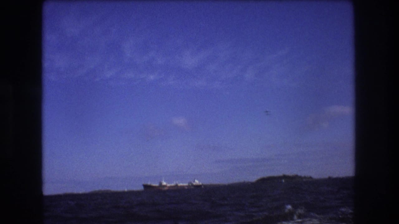 A distant large ship appears on the horizon beyond choppy water under a blue sky, seen from a moving boat in the Boston-area 1975 footage.