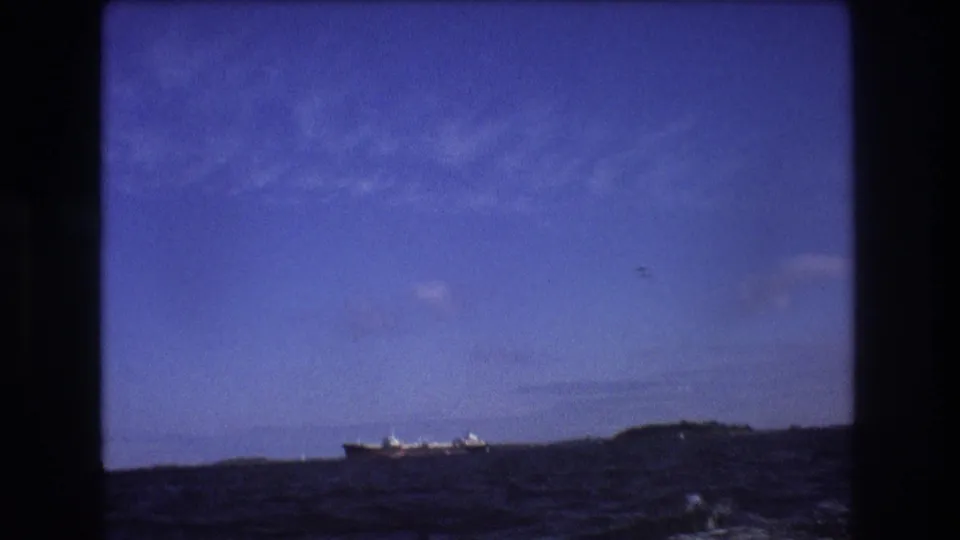 A distant large ship appears on the horizon beyond choppy water under a blue sky, seen from a moving boat in the Boston-area 1975 footage.