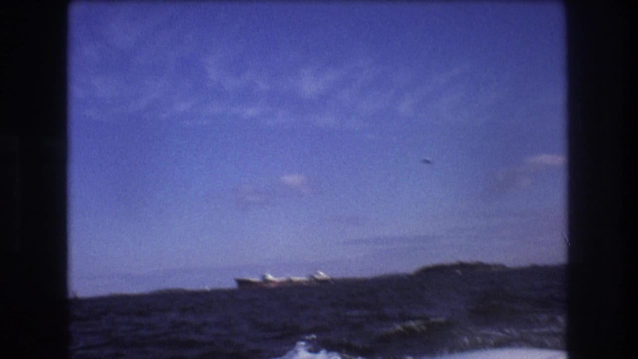 The camera looks across dark, uneven water toward a large ship and low shoreline, with the handheld 8mm frame emphasizing the boat's motion.