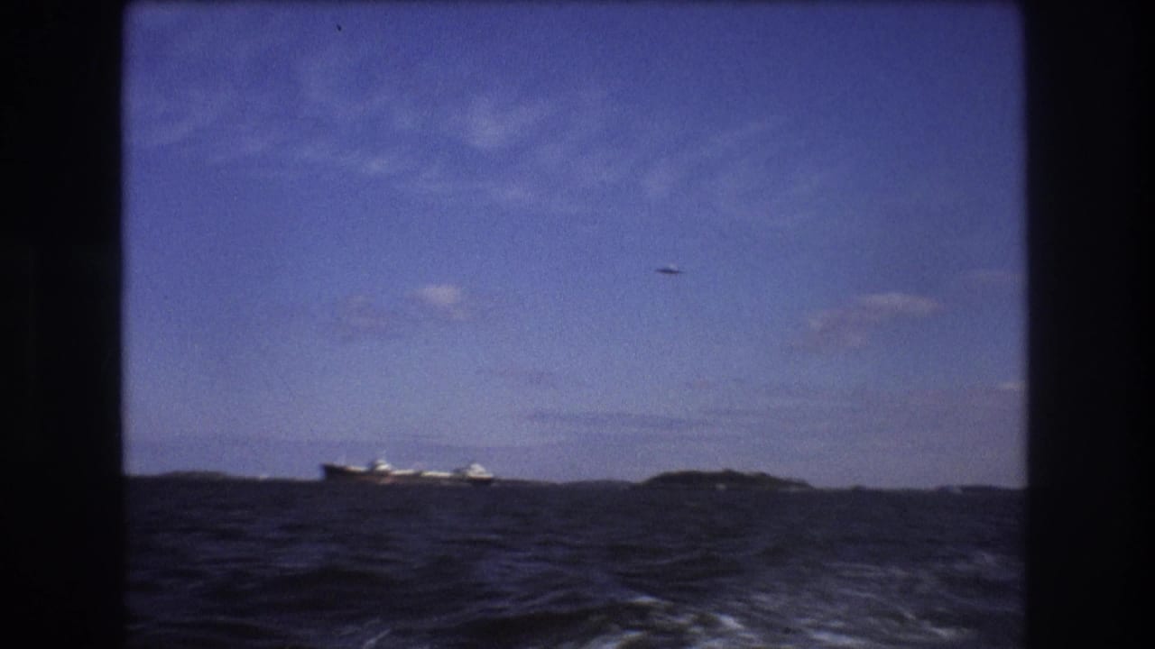 1975 Boston-area 8mm footage shows dark choppy water from a moving motorboat, with a distant cargo ship near the horizon and a small aircraft overhead.