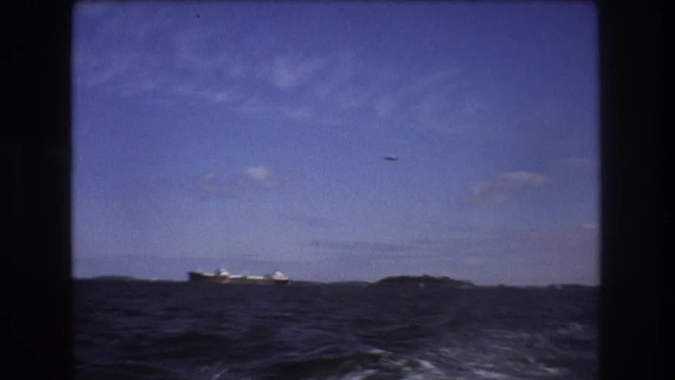 1975 Boston-area 8mm footage shows dark choppy water from a moving motorboat, with a distant cargo ship near the horizon and a small aircraft overhead.