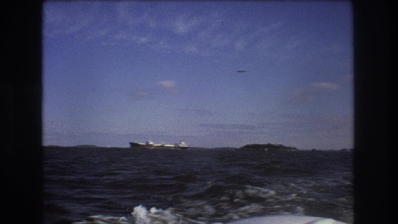 From a moving boat, the frame shows rough coastal water, white wake in the foreground, a distant ship on the horizon, and a small aircraft crossing the blue sky.