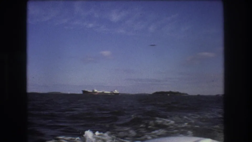 From a moving boat, the frame shows rough coastal water, white wake in the foreground, a distant ship on the horizon, and a small aircraft crossing the blue sky.