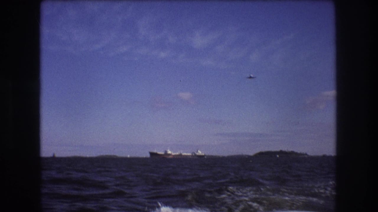The 8mm frame holds on choppy blue water with a distant cargo vessel, low shoreline or island forms, and a small aircraft visible against the sky.