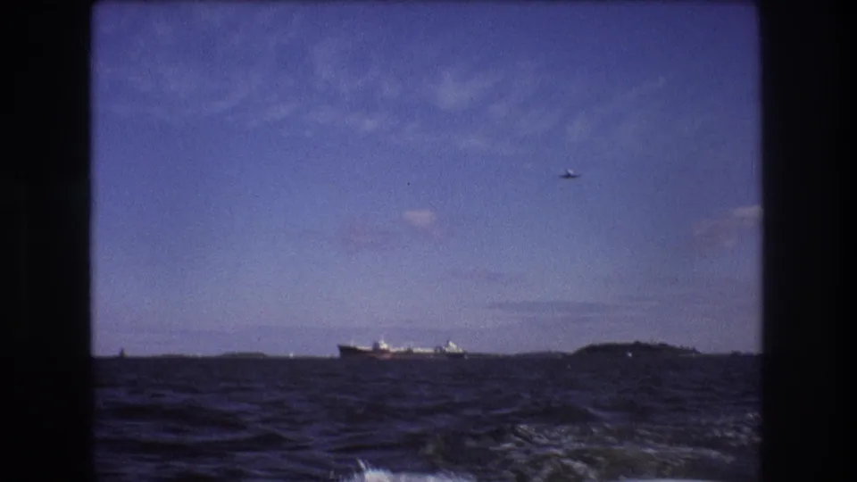 The 8mm frame holds on choppy blue water with a distant cargo vessel, low shoreline or island forms, and a small aircraft visible against the sky.