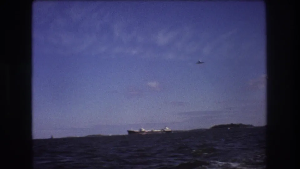 A final 1975 Boston-area boat-view frame shows choppy water, a distant ship beside low landforms, and a small aircraft high in the blue sky.