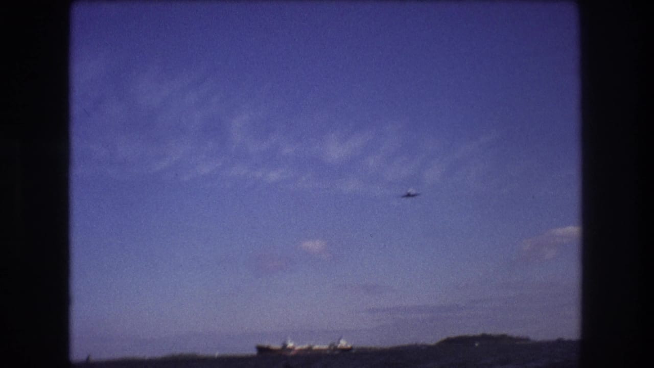 1975 Boston-area 8mm frame from a moving boat showing choppy ocean water, a distant cargo ship near the horizon, low shoreline, and a small airplane against a blue sky.