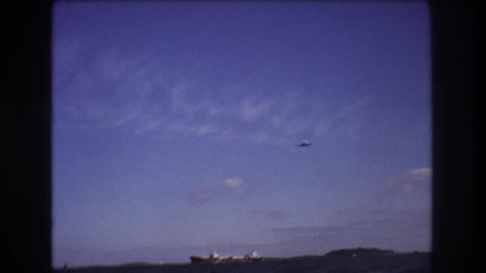 1975 Boston-area 8mm frame from a moving boat showing choppy ocean water, a distant cargo ship near the horizon, low shoreline, and a small airplane against a blue sky.