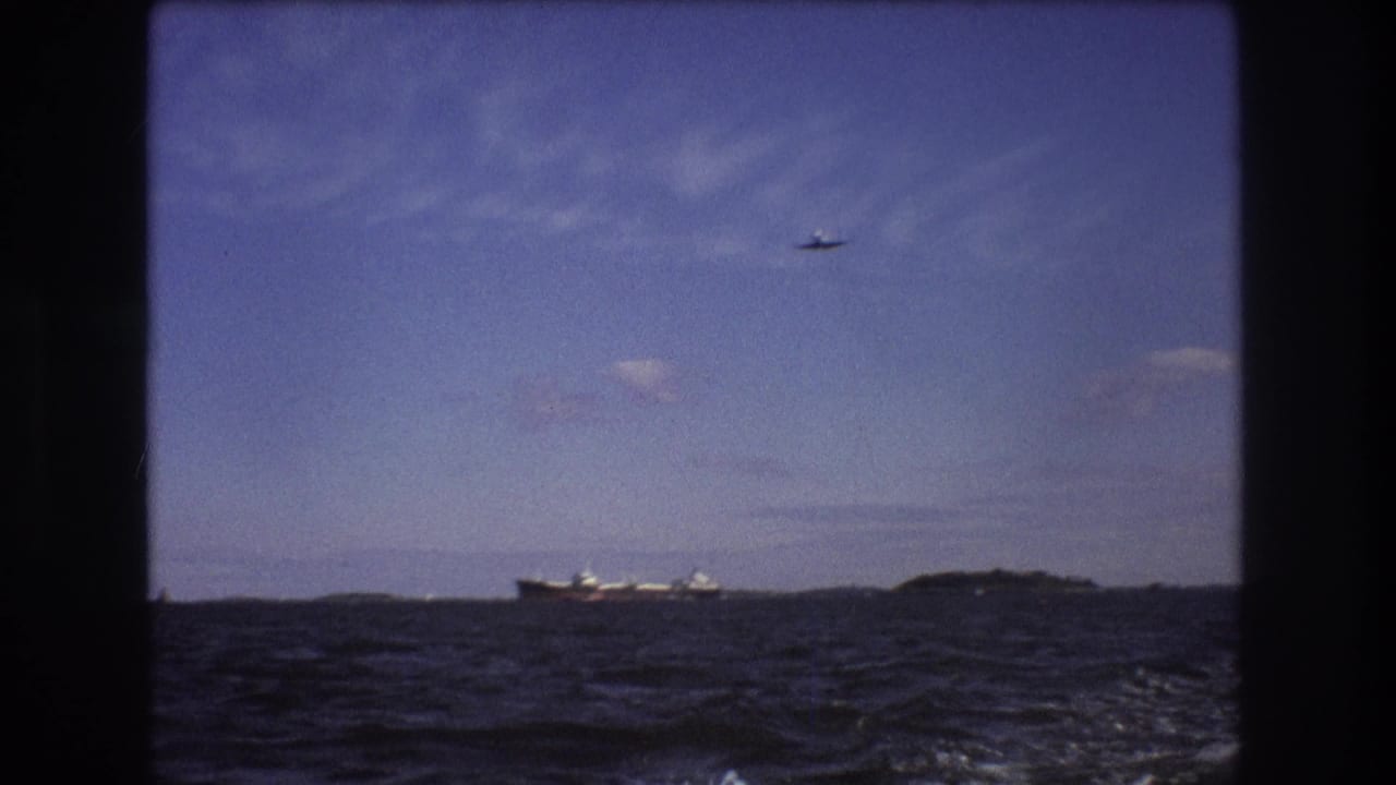 1975 Boston-area 8mm frame showing a distant cargo ship beyond choppy water, a low landform on the horizon, and a small airplane crossing the blue sky.