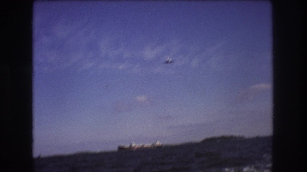 1975 Boston-area 8mm frame with a small airplane overhead, a cargo ship near the horizon, choppy sea in the foreground, and soft clouds across a blue sky.