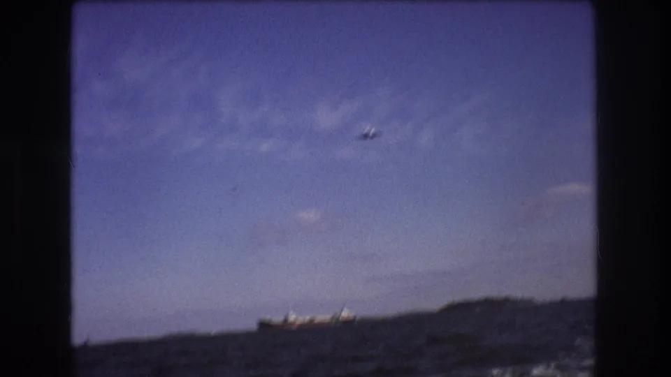1975 Boston-area 8mm frame with a small airplane overhead, a cargo ship near the horizon, choppy sea in the foreground, and soft clouds across a blue sky.
