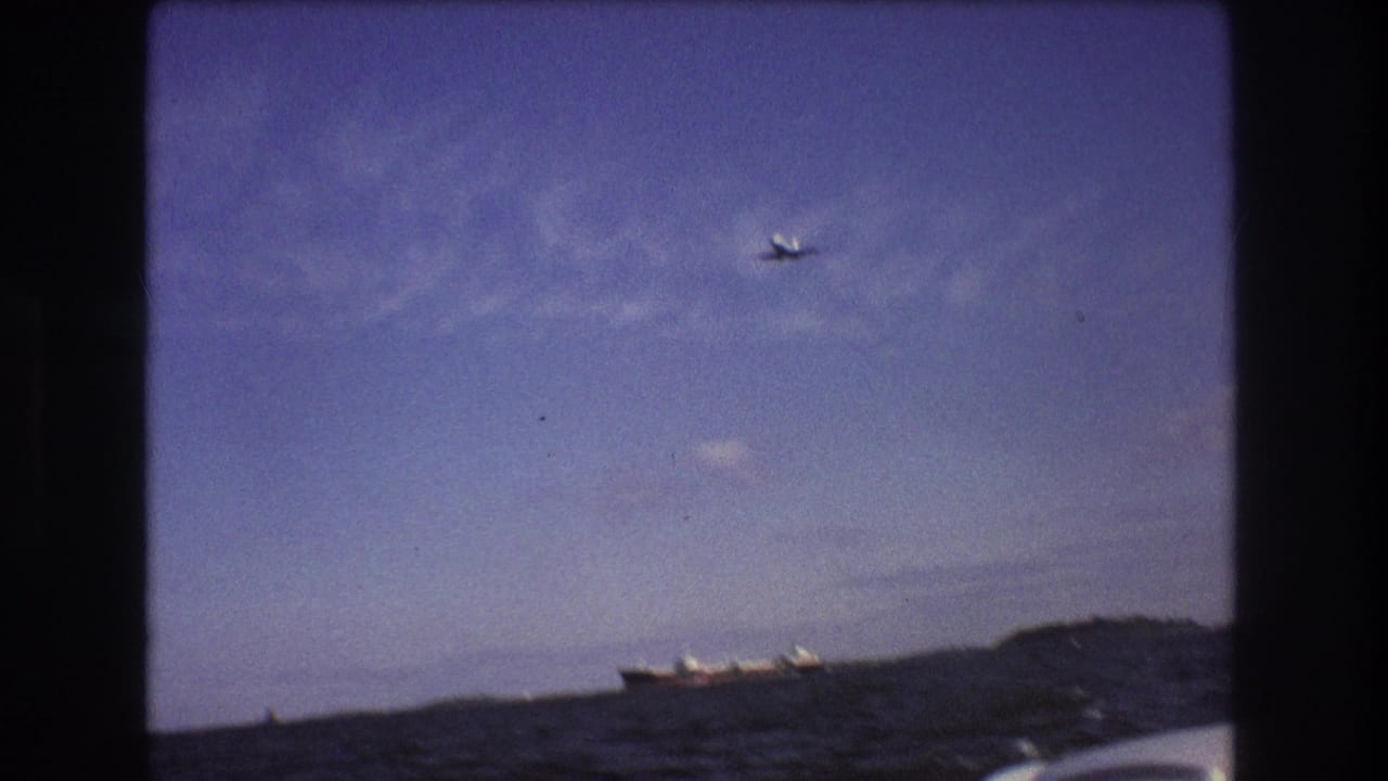 1975 Boston-area 8mm frame from a boat showing choppy ocean water, a distant cargo ship, a small airplane overhead, and a low shoreline under a blue sky.