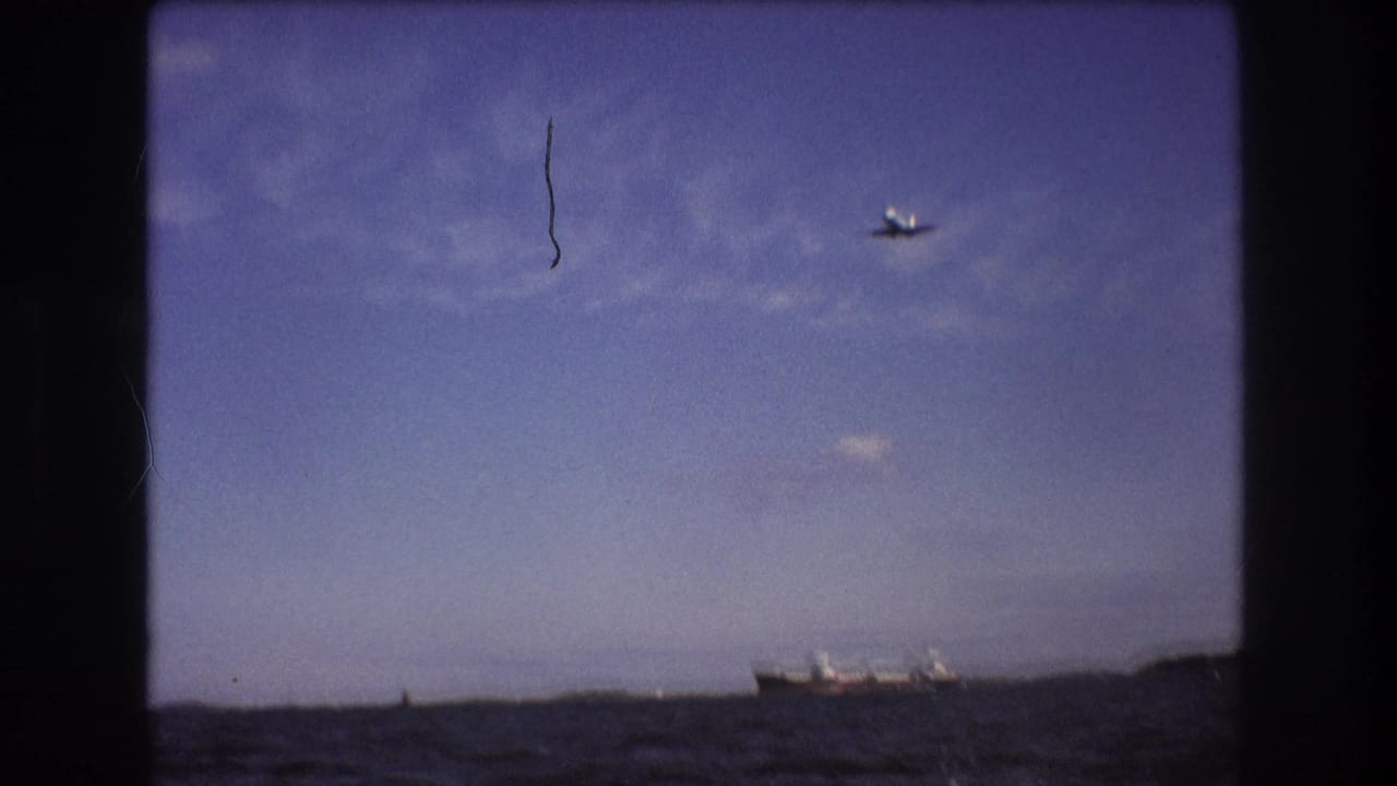 Small commercial airplane seen in flight over distant land or shoreline in supplied 1975 Boston-area 8mm footage, framed by dark film gate edges.