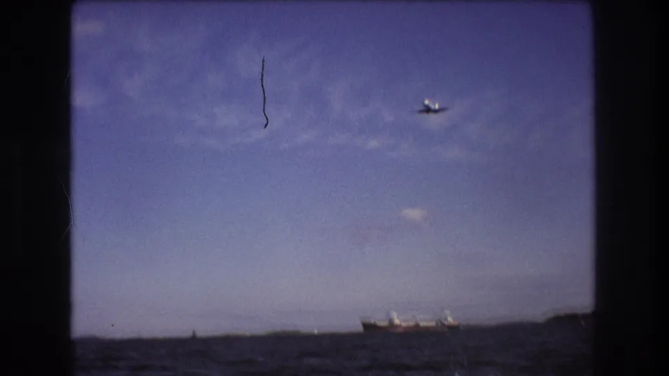Small commercial airplane seen in flight over distant land or shoreline in supplied 1975 Boston-area 8mm footage, framed by dark film gate edges.