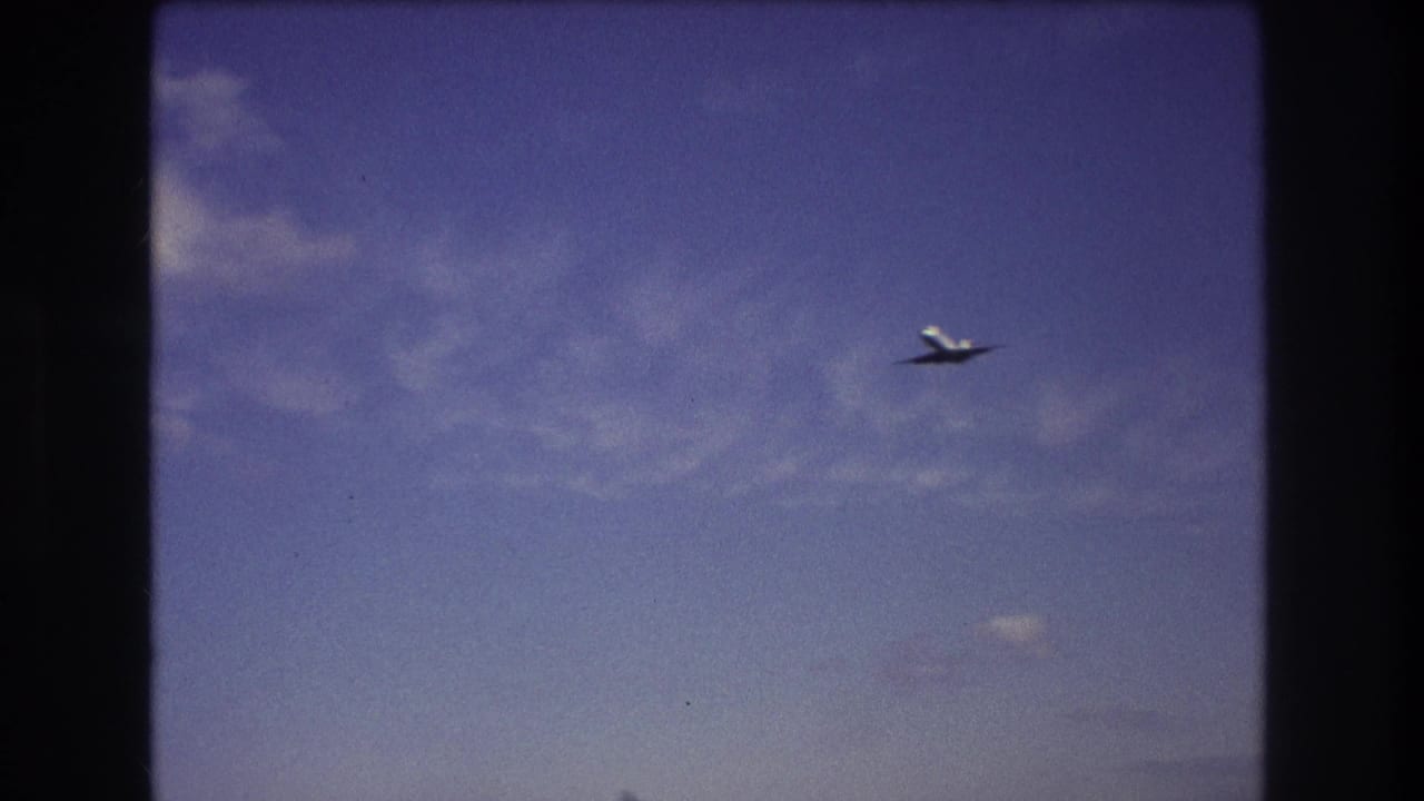 Commercial airplane appears small against a deep blue sky with thin clouds in the supplied 1975 Boston home-movie footage.