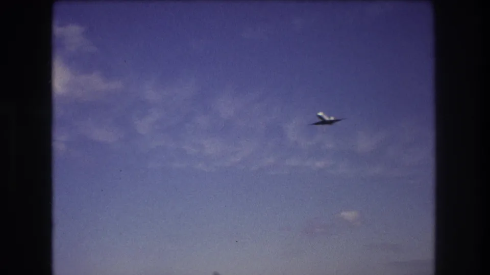 Commercial airplane appears small against a deep blue sky with thin clouds in the supplied 1975 Boston home-movie footage.