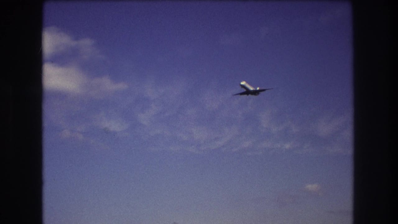 A commercial airplane is framed mid-distance against a blue, lightly clouded sky in 8mm footage identified as Boston in 1975.