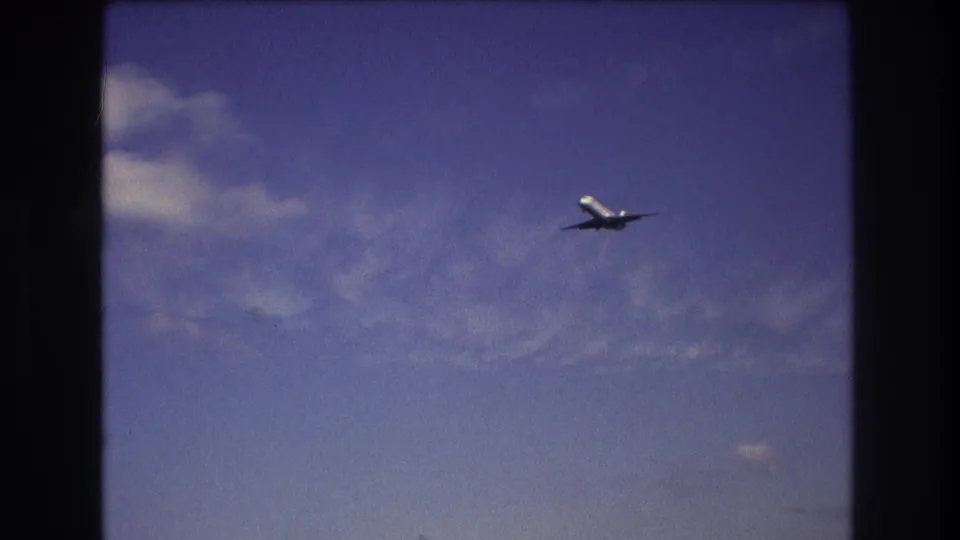 A commercial airplane is framed mid-distance against a blue, lightly clouded sky in 8mm footage identified as Boston in 1975.